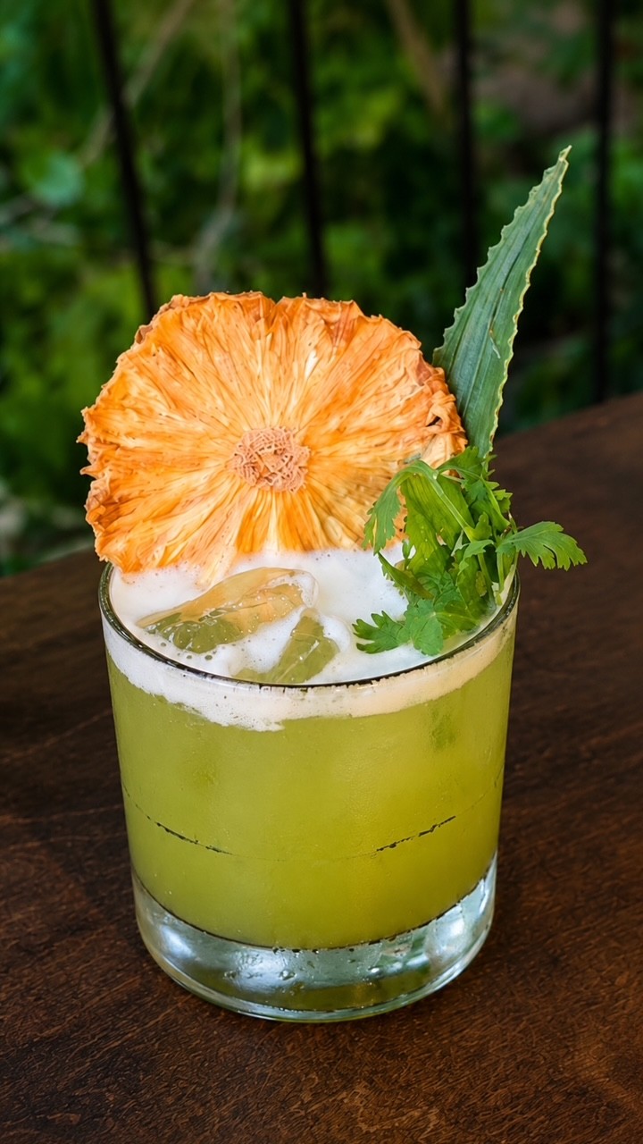 A green cocktail garnished with dried pineapple and fresh cilantro